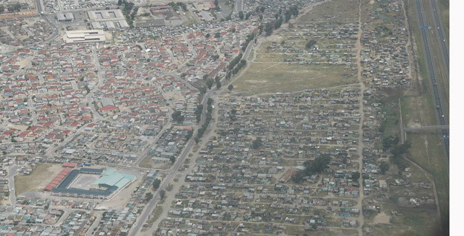 Nyomornegyed Dél-Afrikában: balra a színesbőrűek által lakott, de aránylag rendezett lakónegyed, majd az út túloldalán a kép közepén a valódi bádogváros. Az autópálya a kép jobb oldalán nem csak határként működik, hanem igazodási pont is: a slum lakói ennek mentén gyalogolnak a belső kerültek felé valami megélhetés után nézni. (Trócsányi A. felvétele)